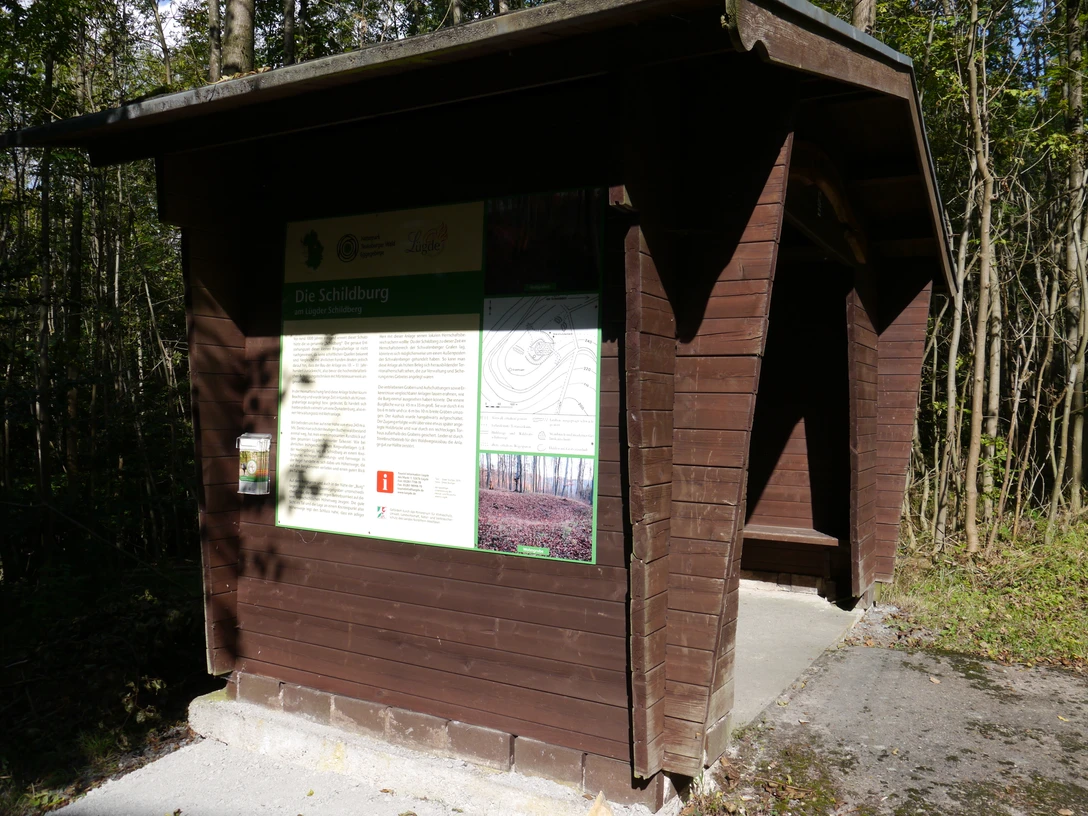 Schutzhütte mit Infotafel Hölzerne Schutzhütte im Wald mit Infotafel zur Schildburg, umgeben von Bäumen und Sonnenlicht.