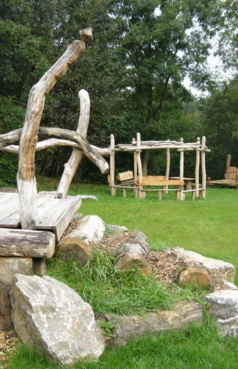 Rast- und Spielplatz Almena Holzspielgeräte und eine überdachte Sitzgelegenheit auf einer Wiese, umringt von Bäumen.