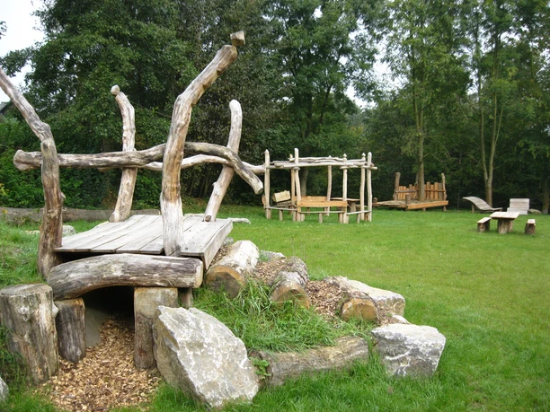 Rast- und Spielplatz Almena Holzspielgeräte und eine überdachte Sitzgelegenheit auf einer Wiese, umringt von Bäumen.