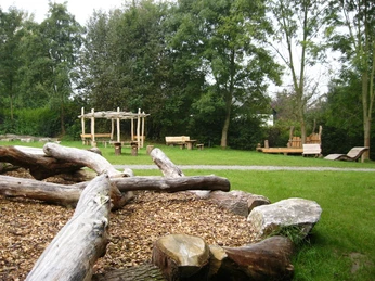 Rast- und Spielplatz Almena Rast- und Spielplatz mit Holzklettergerüst, Bänken und viel Grünfläche, umgeben von hohen Bäumen.