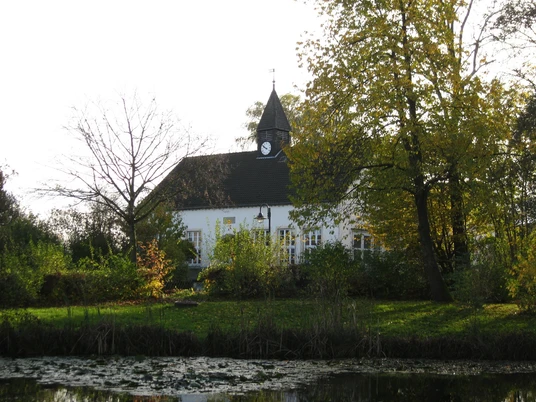 Alte Schule Laßbruch Historisches Gebäude mit Turm, umgeben von Bäumen und einem kleinen Teich in ländlicher Umgebung.