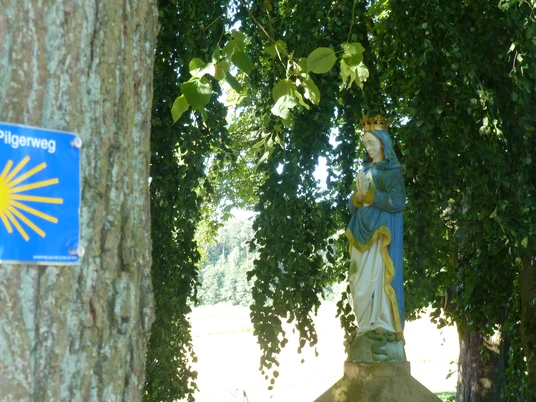 Stehende Marienstatue neben einem Wegweiser mit Pilgermuschel, umgeben von üppigem Grün im Haxtergrund.