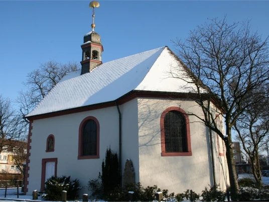 Schneebedeckte kleine Kapelle mit rotem Rahmen an Fenstern und Tür; umgeben von kahlen Bäumen.