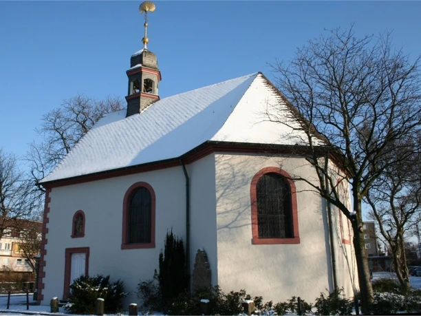 Schneebedeckte kleine Kapelle mit rotem Rahmen an Fenstern und Tür; umgeben von kahlen Bäumen.