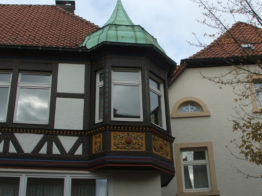 Fachwerkgebäude mit Erker und grünem Kupferdach vor bewölktem Himmel, rechts angrenzendes Gebäude.