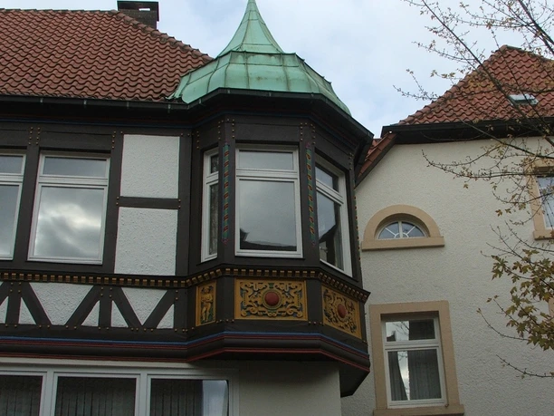 Fachwerkgebäude mit Erker und grünem Kupferdach vor bewölktem Himmel, rechts angrenzendes Gebäude.