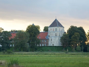 Historisches Schloss mit rotem Dach, umgeben von Bäumen und grünen Wiesen in der Dämmerung.