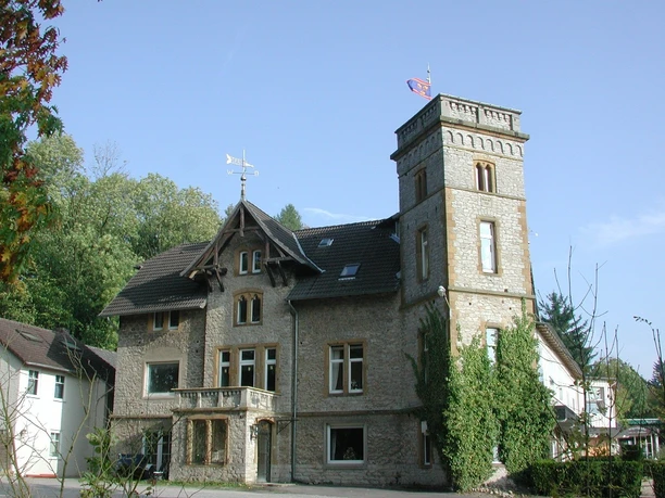 Historische Steinvilla mit Turm, umgeben von Bäumen; blauer Himmel und wehende Fahne im Hintergrund.