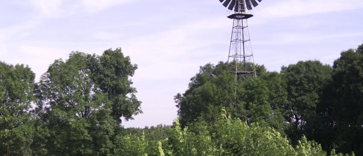 Windrad Ein Windrad erhebt sich über Bäumen, flankiert von einem Feld und einem Holzzaun unter blauem Himmel.