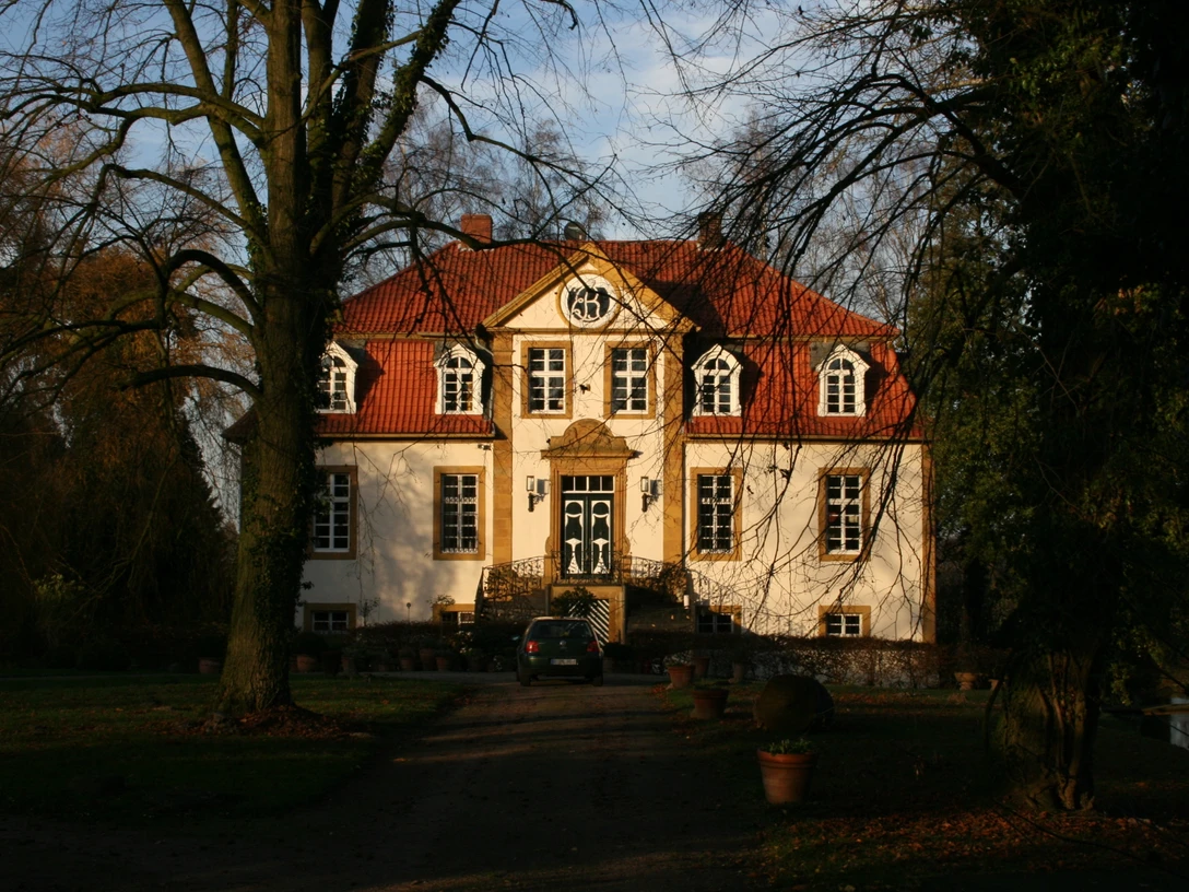 Herrenhaus mit rotem Dach und weißen Fassaden, umgeben von Bäumen, bei tiefstehender Sonne.