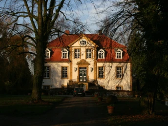Herrenhaus mit rotem Dach und weißen Fassaden, umgeben von Bäumen, bei tiefstehender Sonne.