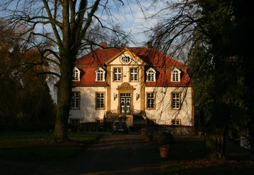 Haus Bosfeld Herrenhaus mit rotem Dach und weißen Fassaden, umgeben von Bäumen, bei tiefstehender Sonne.