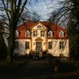 Haus Bosfeld Herrenhaus mit rotem Dach und weißen Fassaden, umgeben von Bäumen, bei tiefstehender Sonne.