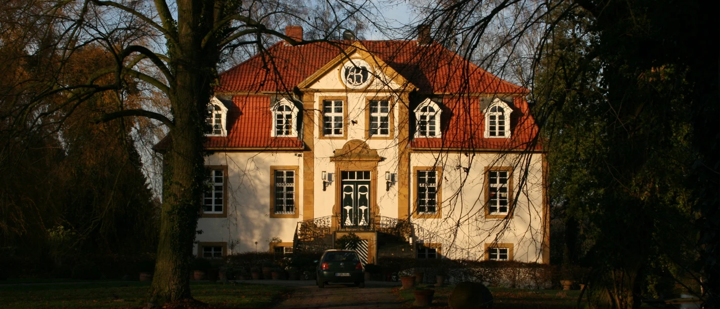 Haus Bosfeld Herrenhaus mit rotem Dach und weißen Fassaden, umgeben von Bäumen, bei tiefstehender Sonne.