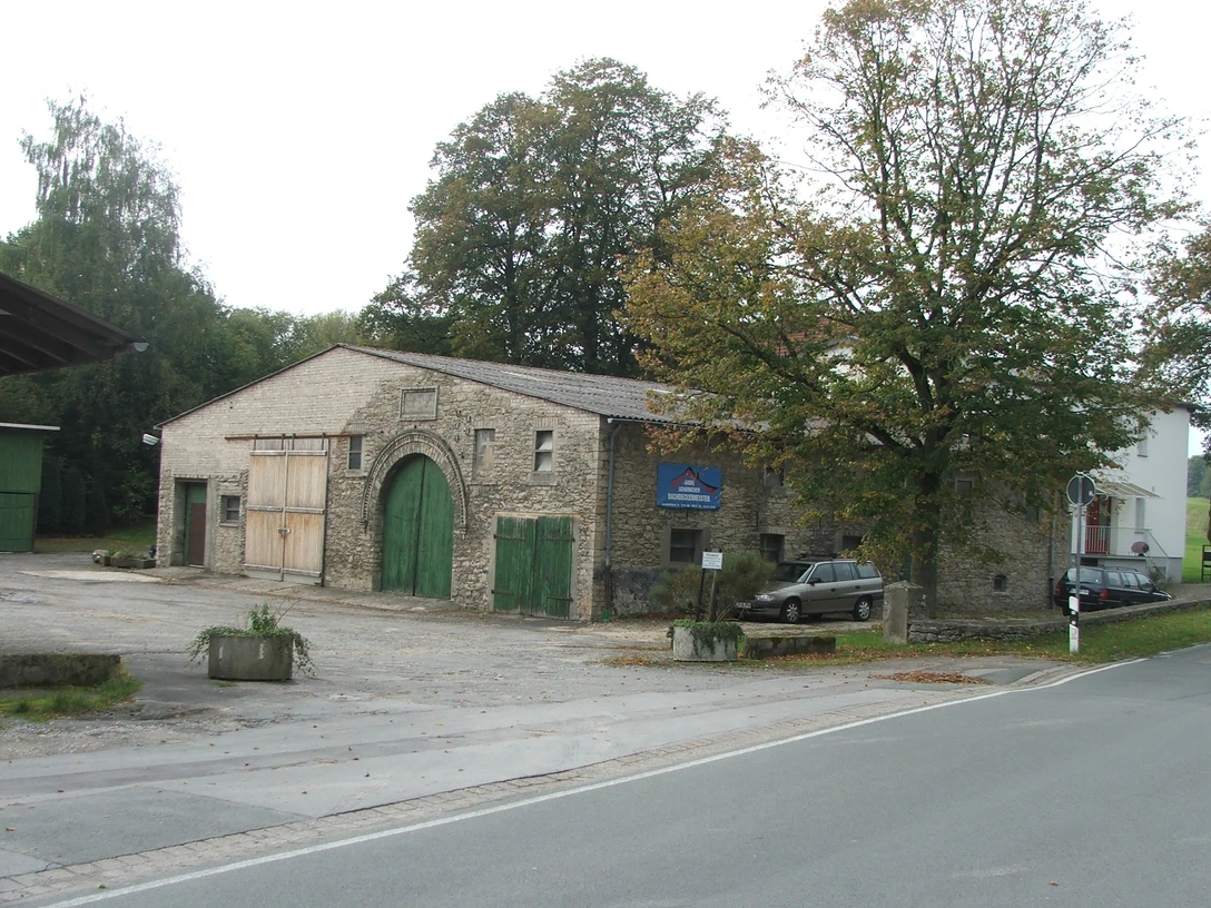 Steinernes Bauernhaus mit großen Toren, von Bäumen umgeben, an einer ruhigen Landstraße gelegen.