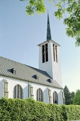 Eine weiße Kirche mit steilem Turm und Spitzdach, umgeben von Bäumen unter klarem Himmel.