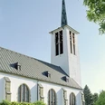 Eine weiße Kirche mit steilem Turm und Spitzdach, umgeben von Bäumen unter klarem Himmel.