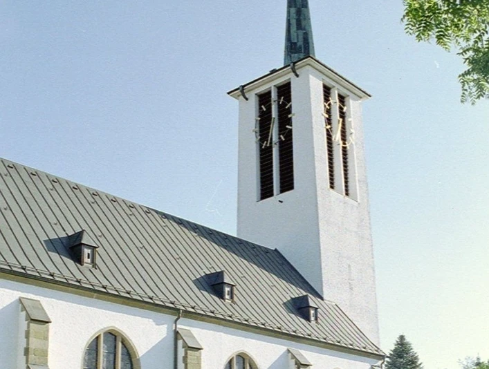 Eine weiße Kirche mit steilem Turm und Spitzdach, umgeben von Bäumen unter klarem Himmel.