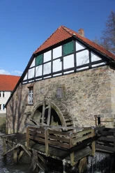 Ölmühle Historische Fachwerk-Ölmühle mit rotem Ziegeldach und großem hölzernem Wasserrad an einem klaren Bach.