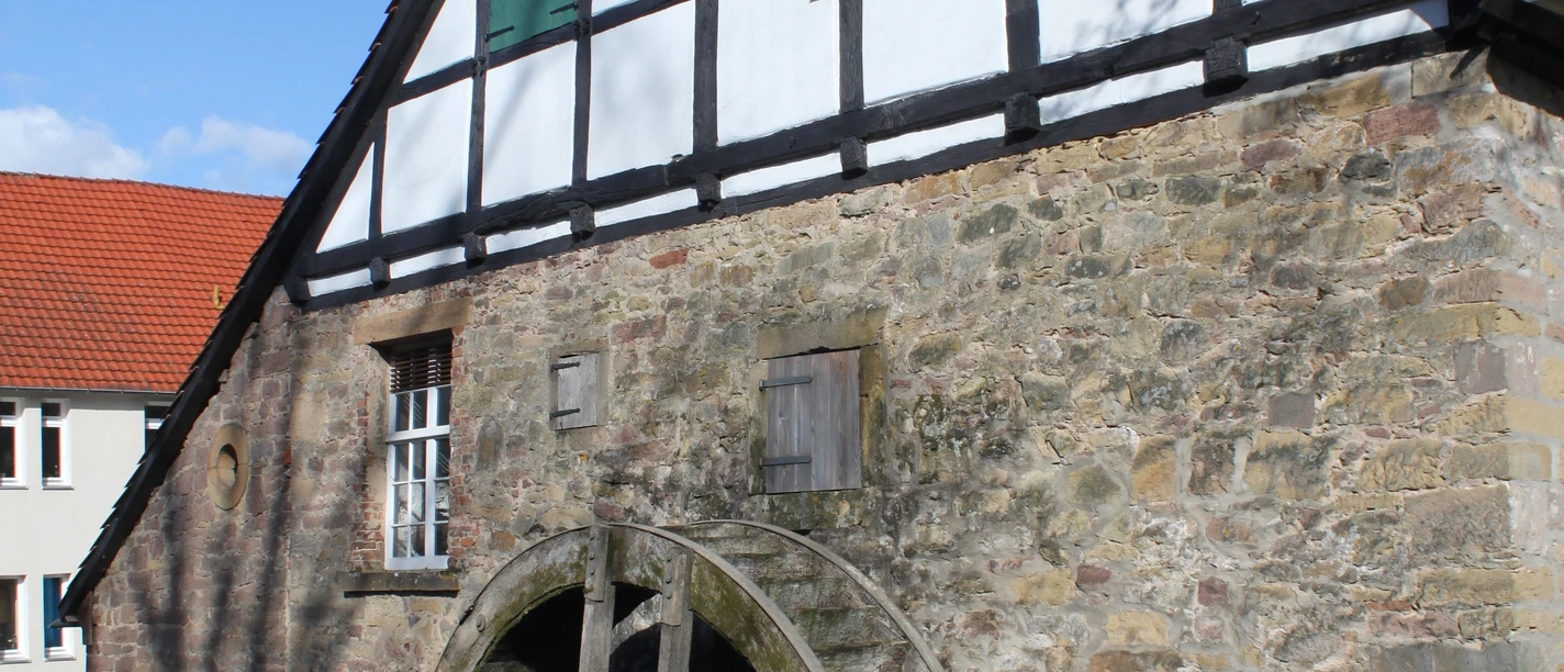 Ölmühle Historische Fachwerk-Ölmühle mit rotem Ziegeldach und großem hölzernem Wasserrad an einem klaren Bach.