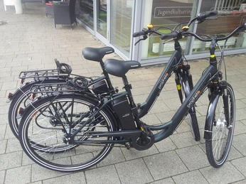 Pedelecs Drei schwarze Elektrofahrräder stehen nebeneinander auf einem gepflasterten Gehweg vor einem Gebäude.