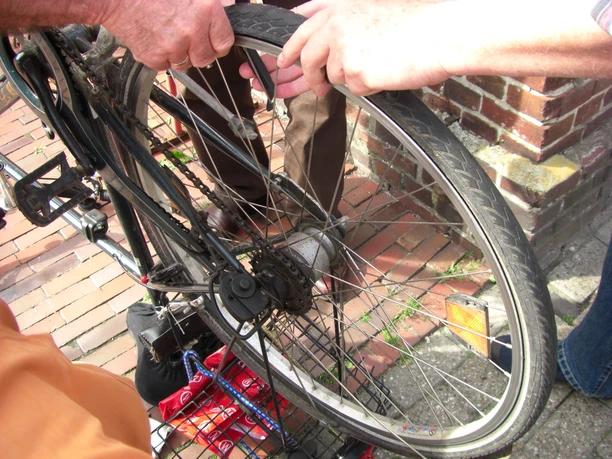Fahrradfachgeschäft Zwei Personen reparieren gemeinsam ein Fahrradlaufrad auf einem Pflasterweg neben einer Backsteinmauer.