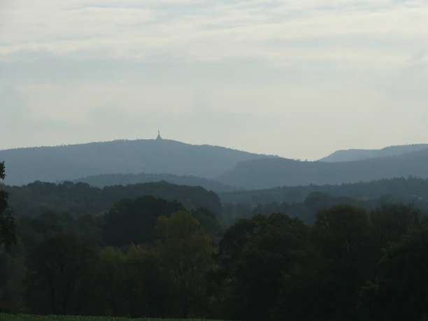 Hügelige Landschaft mit dichtem Wald, im Hintergrund ein Monument auf einem entfernten Bergkamm.