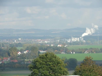 Blick nach Lage Hügelige Landschaft mit Feldern und Bäumen, im Hintergrund eine Stadt und ein Industriekomplex.