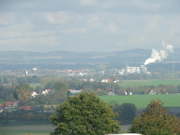 Blick nach Lage Hügelige Landschaft mit Feldern und Bäumen, im Hintergrund eine Stadt und ein Industriekomplex.