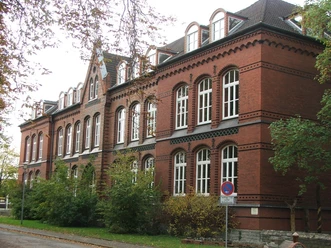 Backsteinbau der Bürgerschule mit vielen Fenstern und Dachgauben, umgeben von Bäumen und Herbstlaub.