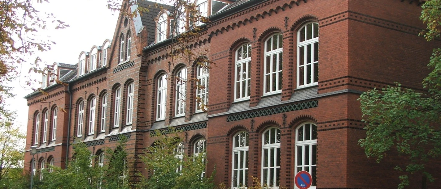 Backsteinbau der Bürgerschule mit vielen Fenstern und Dachgauben, umgeben von Bäumen und Herbstlaub.