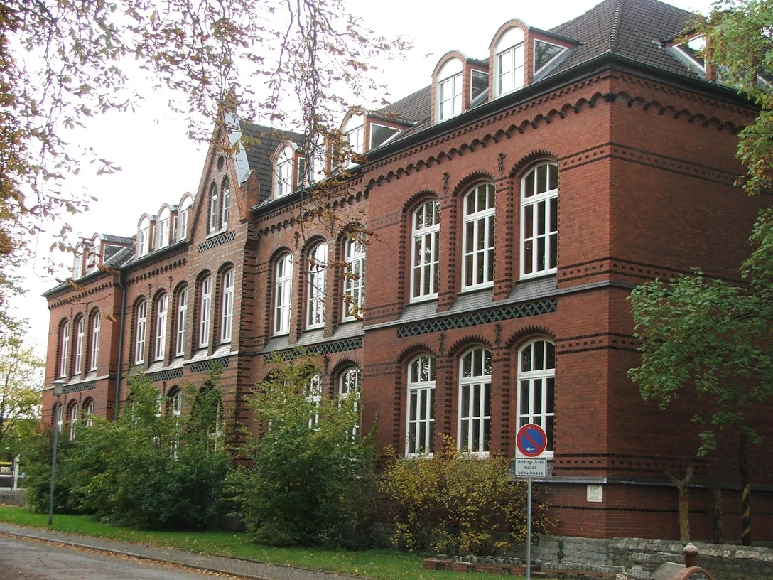 Bürgerschule Backsteinbau der Bürgerschule mit vielen Fenstern und Dachgauben, umgeben von Bäumen und Herbstlaub.