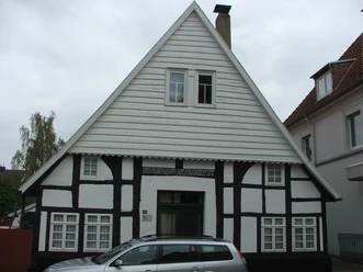 Fachwerkhaus mit weiß-schwarzer Fassade und steilem Giebel, geparktes Auto davor, bewölkter Himmel.