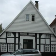 Fachwerkhaus Rhienstraße 43 Fachwerkhaus mit weiß-schwarzer Fassade und steilem Giebel, geparktes Auto davor, bewölkter Himmel.
