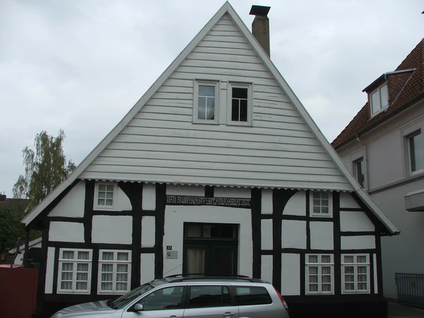 Fachwerkhaus Rhienstraße 43 Fachwerkhaus mit weiß-schwarzer Fassade und steilem Giebel, geparktes Auto davor, bewölkter Himmel.