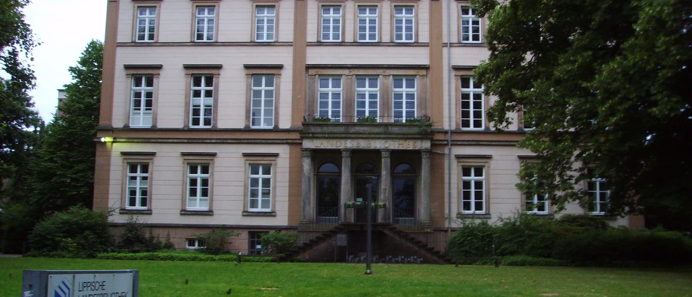 Lippische Landesbibliothek Historisches, helles Gebäude mit roten Dachziegeln, großen Fenstern und einer gepflegten Rasenfläche.