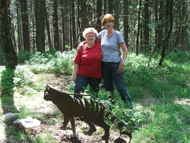 Ortrud Hinze, Karen Tank Zwei Frauen stehen nahe einer Metallskulptur eines Tigers in einem sonnigen Waldstück.