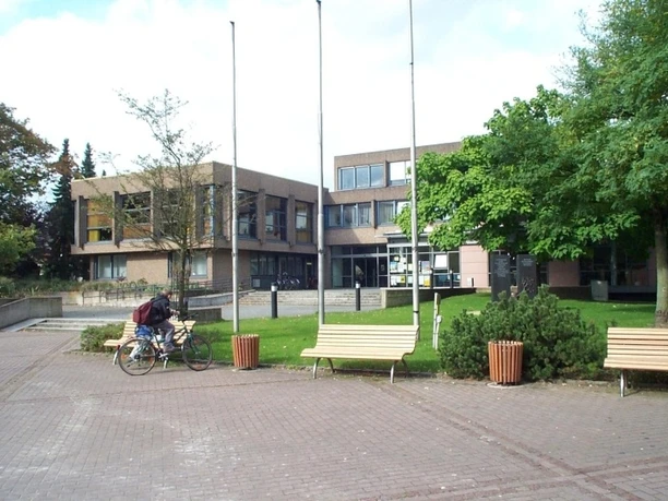 Touristinfo im Rathaus Versmold Ziegelgebäude mit modernen Fenstern, drei Fahnenmasten und grünen Bäumen, davor eine Person mit Fahrrad.