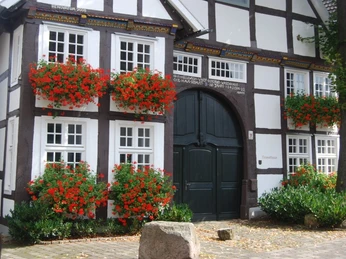 Heimathaus Rietberg Fachwerkhaus mit blumengeschmückten Fenstern, dunklem Holztor und weißen Wänden in ländlicher Umgebung.