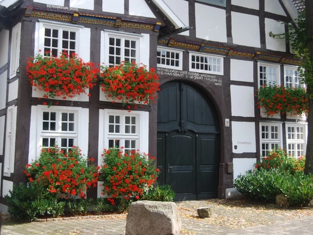 Heimathaus Rietberg Fachwerkhaus mit blumengeschmückten Fenstern, dunklem Holztor und weißen Wänden in ländlicher Umgebung.