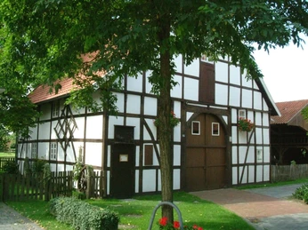 Fachwerkhaus mit rotem Dach, umgeben von grünem Rasen und großen Bäumen an einem sonnigen Tag.