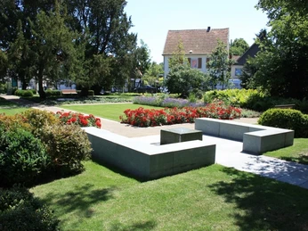 Ein gepflegter Park mit grüner Wiese, gepflastertem Weg, roten Blumenbeeten und modernen Betonbänken.