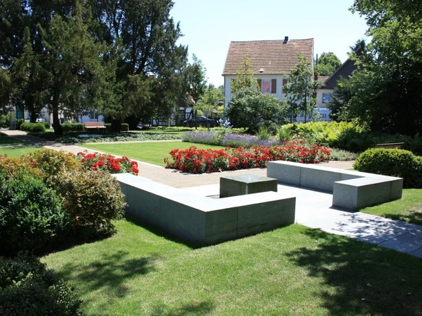 Ein gepflegter Park mit grüner Wiese, gepflastertem Weg, roten Blumenbeeten und modernen Betonbänken.