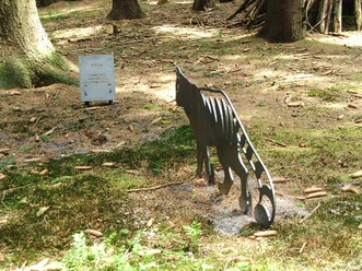 Kitty come back Stählerne Katzenfigur in einem lichtdurchfluteten Waldstück neben einer Informationstafel.