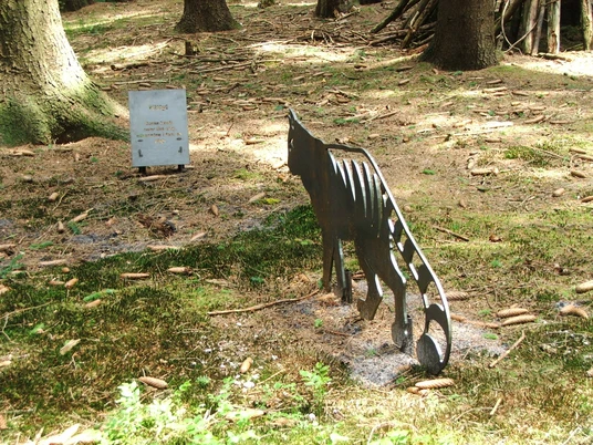 Kitty come back Stählerne Katzenfigur in einem lichtdurchfluteten Waldstück neben einer Informationstafel.