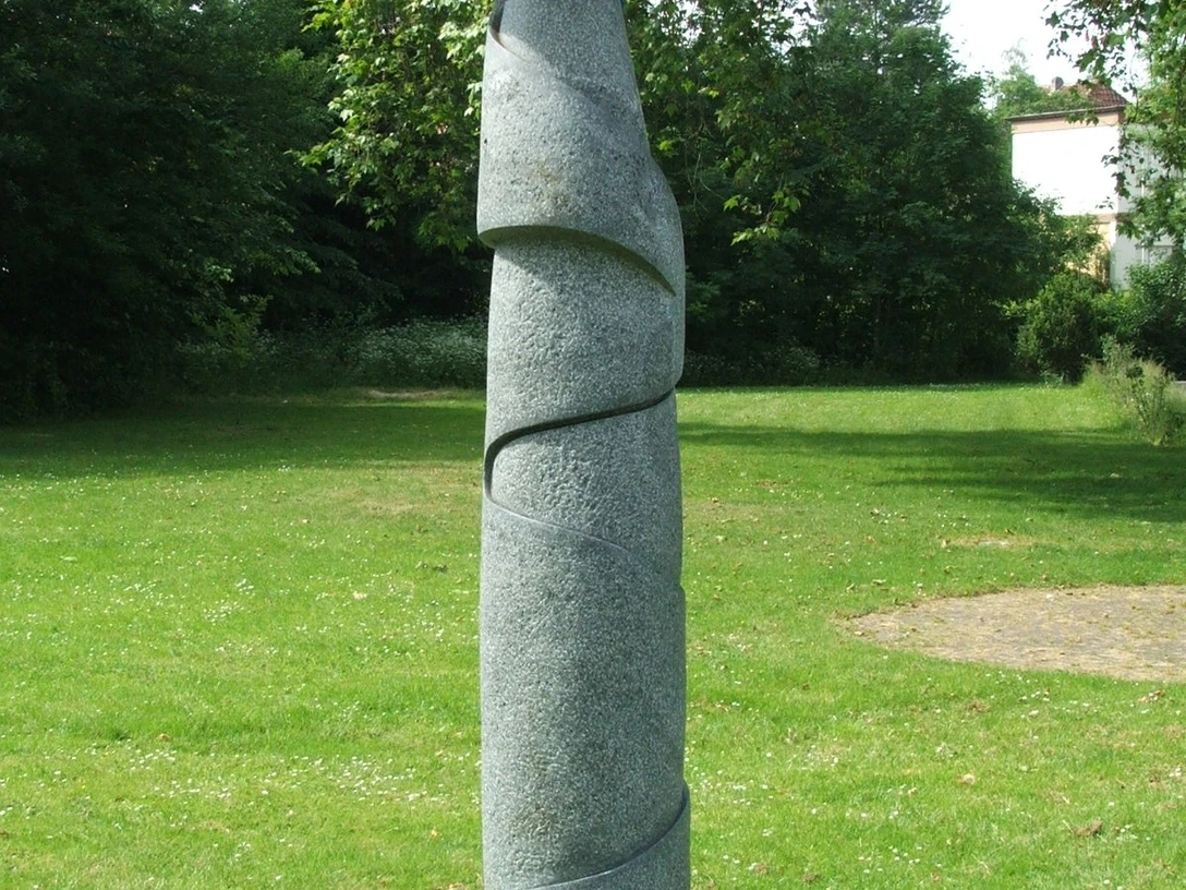 Moderne Skulptur auf Rasenfläche, bestehend aus spiralförmiger Steinstruktur mit blauer Metallplatte oben.