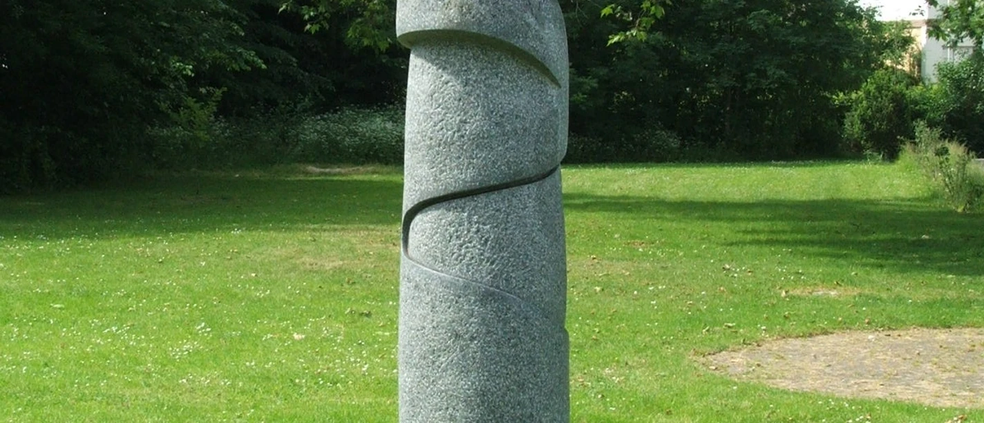 Moderne Skulptur auf Rasenfläche, bestehend aus spiralförmiger Steinstruktur mit blauer Metallplatte oben.