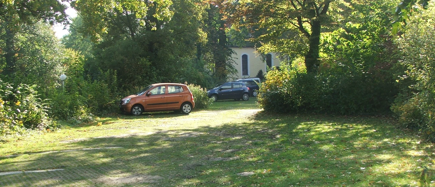 Parkplatz mit zwei Autos vor einer von Bäumen umgebenen Kirche, darunter ein rotes und ein blaues Fahrzeug.