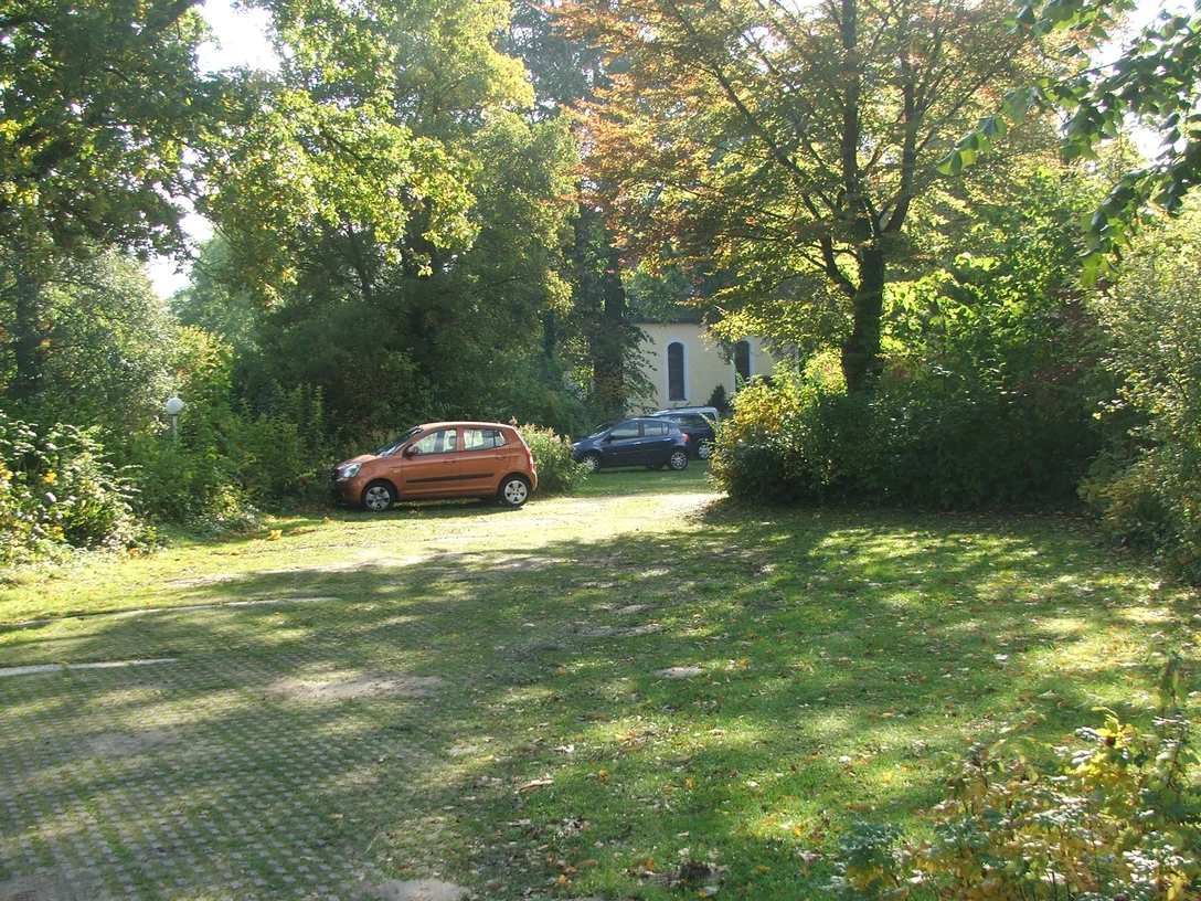 Parkplatz Kirche Stapelage Parkplatz mit zwei Autos vor einer von Bäumen umgebenen Kirche, darunter ein rotes und ein blaues Fahrzeug.