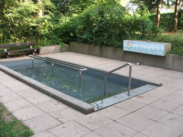 Kneipptretbecken Kneipptretbecken umgeben von Bäumen, mit Haltestangen und Steinboden für eine erfrischende Pause.